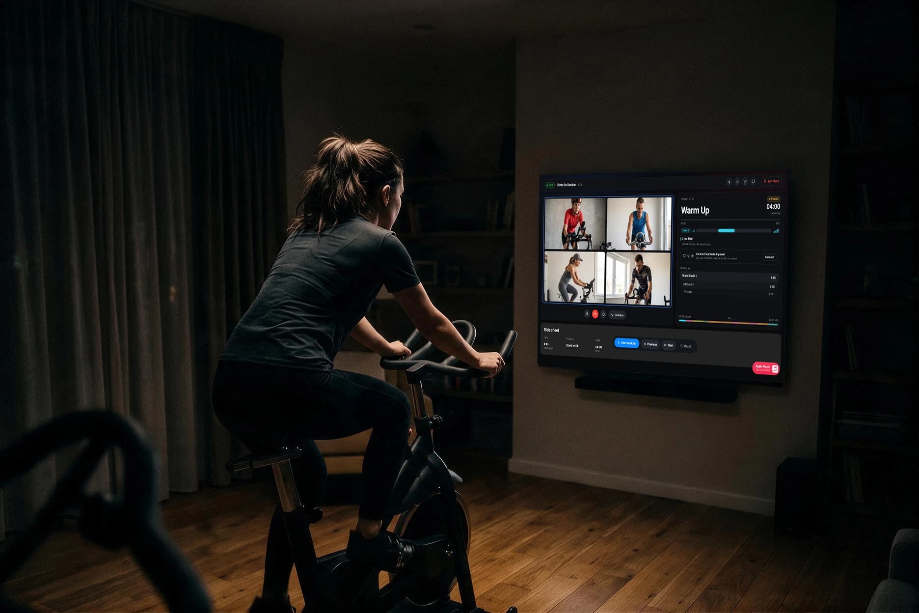 A rider on a stationary bike in a dark living room, facing a wall-mounted screen showing a live Ride Director class: four riders on video and the current stage counting down
